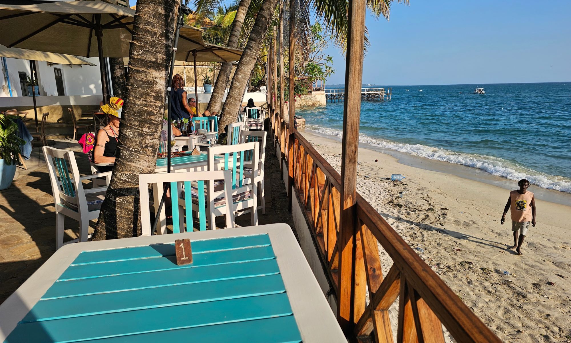 A wooden deck with bar stools under palm trees, facing the sea